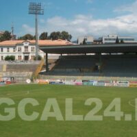 Stadio di calcio all'aperto con spalti, campo verde e strutture circostanti in giornata soleggiata.