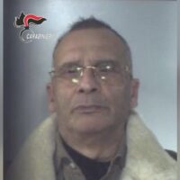 Uomo anziano con occhiali, giacca con ovatta, espressione neutra, sfondo sfumato grigio.
