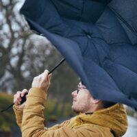 Uomo con cappotto giallo tiene in alto un grande ombrello blu, senza pioggia ma sotto cielo nuvoloso.