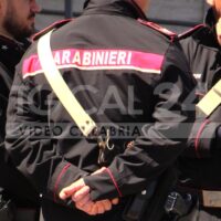 Due carabinieri in uniforme nero con strisce rosse e gialle, mano destra legata dietro, in primo piano.