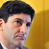 Primo piano di un uomo italiano con espressione seria, sfondo giallo sfumato.