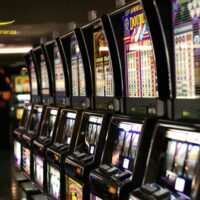 Fila di slot machine luminose in una sala da gioco, luci colorate, schermi e pulsanti, atmosfera vivace e affollata.