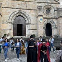 Gruppo di persone in abiti storici davanti a una chiesa in pietra, con archi e facciate antiche sullo sfondo.