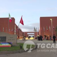 Vista di un campus o istituto con edifici rossi, bandiere e CIP/CIO in primo piano, ingresso centrale e persone ai lati.